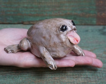 Könnte beinhalten: Eine Keramik-Froschfigur mit einem großen, runden Körper und einem weit geöffneten Maul. Der Frosch hat große, schwarze Augen und eine rosa Zunge, die herausstreckt. Der Frosch ist in Braun- und Grautönen bemalt.