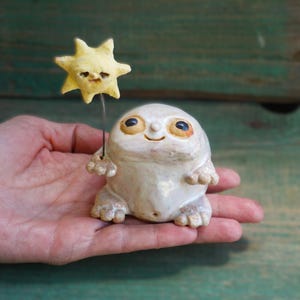 Może przedstawiać: Kapryśna, kremowa figurka ceramiczna stworzenia z dużymi oczami i uśmiechniętą miną, trzymająca żółtą gwiazdę na druciku. Figurka trzymana jest w dłoni na tle zielonego drewnianego tła.