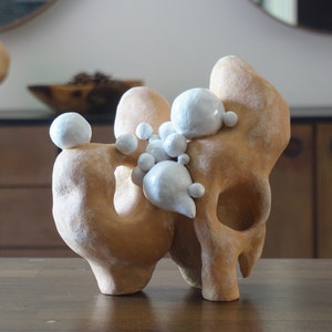 May include: A tan ceramic sculpture with a biomorphic form, featuring rounded shapes and a series of white, spherical accents. The sculpture has a textured surface and a hollowed-out section, suggesting an abstract design.