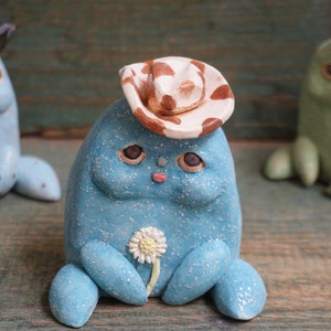 May include: Three ceramic figurines painted in blue, green, and white. Each figurine wears a different hat: a crown, a cowboy hat, and a red mushroom. The figurines have large eyes and a whimsical expression.