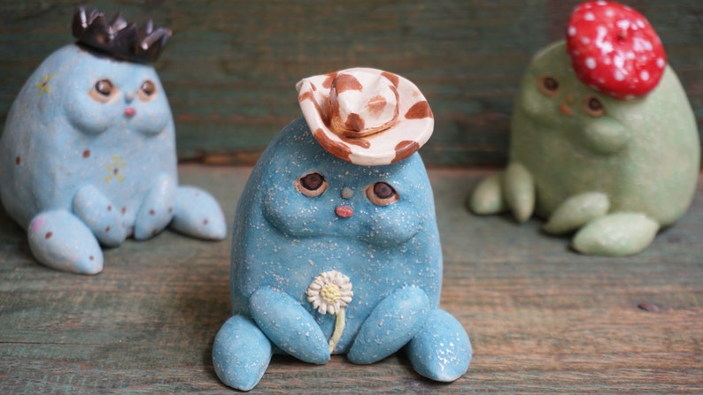 May include: Three ceramic figurines painted in blue, green, and white. Each figurine wears a different hat: a crown, a cowboy hat, and a red mushroom. The figurines have large eyes and a whimsical expression.