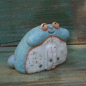 May include: A ceramic figurine of a blue and white creature with large eyes and a wide smile. The creature has a speckled white body with blue accents and a blue head.