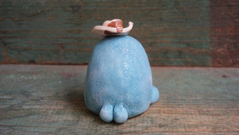 May include: A blue ceramic figurine wearing a white and brown cowboy hat. The figurine has a textured surface and three small feet.