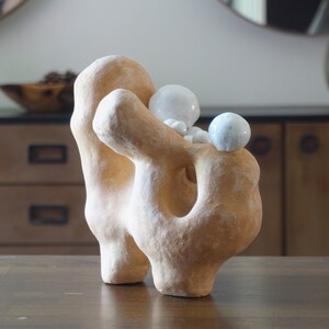 May include: A beige abstract sculpture with a textured surface and a large hole. Several small, white, spherical objects are nestled on top. The sculpture is displayed on a dark wooden table, showcasing its unique form and design.