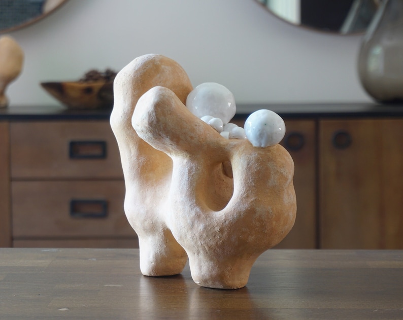 May include: A beige abstract sculpture with a textured surface and a large hole. Several small, white, spherical objects are nestled on top. The sculpture is displayed on a dark wooden table, showcasing its unique form and design.