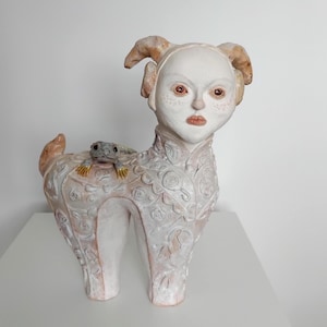 May include: White ceramic sculpture of a stylised animal with a human-like face and horns. The animal is decorated with a floral pattern and has a small grey lizard on its back.