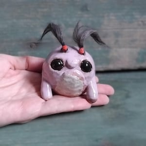 May include: A quirky, lilac-hued ceramic figurine with big, black eyes and two tufts of grey hair secured with red bands. The figure has a rounded body and small legs, held in a hand against a blurred background.