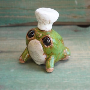 Könnte beinhalten: Eine kleine, runde, grüne Keramikfroschfigur mit glänzender Oberfläche, großen, dunklen, reflektierenden Augen und einem weißen Bauch. Der Frosch trägt eine weiße, texturierte Kochmütze auf dem Kopf und hat zwei Vorderbeine nach vorne gestreckt.