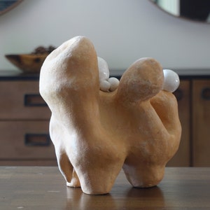 May include: An abstract ceramic sculpture in a tan hue with a textured finish. The sculpture features rounded shapes and is decorated with small, white, spherical details. It is presented on a wooden surface.