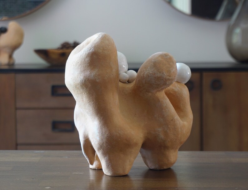 May include: An abstract ceramic sculpture in a tan hue with a textured finish. The sculpture features rounded shapes and is decorated with small, white, spherical details. It is presented on a wooden surface.
