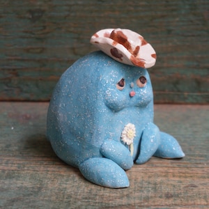 May include: A small, blue, ceramic figurine wearing a brown and white cowboy hat. The figurine has a flower in its hand and a sad expression on its face.