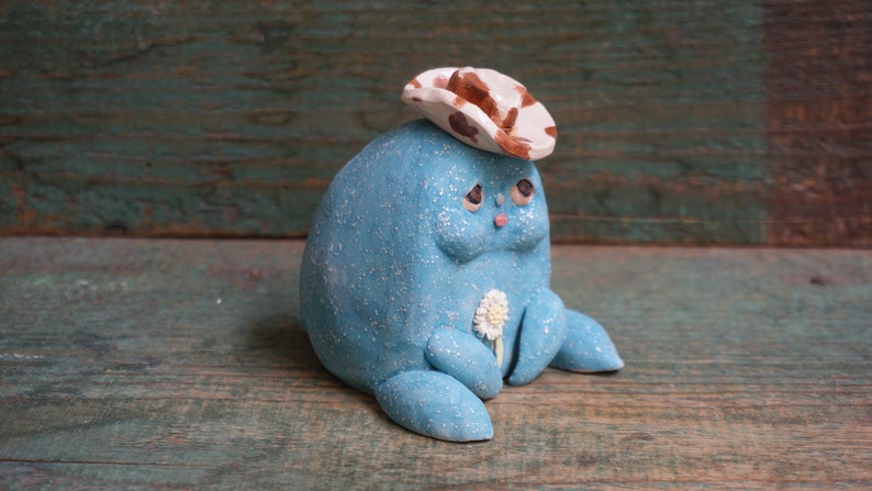 May include: A small, blue, ceramic figurine wearing a brown and white cowboy hat. The figurine has a flower in its hand and a sad expression on its face.