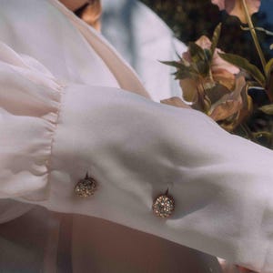 May include: Close-up of a pale pink long-sleeved blouse with two decorative, round, jewelled buttons on the cuff. The blouse is made of a soft, flowing fabric. Flowers with pink petals are in the background.