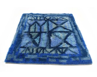 Puede incluir: Una alfombra cuadrada y peluda en tonos azules. La alfombra presenta un diseño geométrico en azul más oscuro, con un fondo azul claro y gris. El borde es azul oscuro con un patrón de cuadros. La alfombra está hecha de un material suave y afelpado.