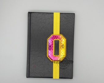 May include: A black notebook with a yellow bookmark and a pink and yellow letter O on the front.