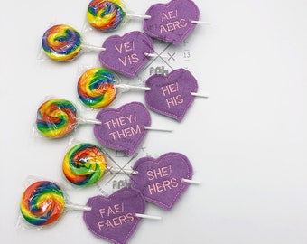 May include: Rainbow swirl lollipops in clear wrappers are displayed with purple heart-shaped felt ornaments. The ornaments have embroidered text including "AE/AERS", "VE/VIS", "HE/HIS", "THEY/THEM", "SHE/HERS", and "FAE/FAERS".