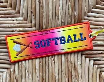 May include: A colorful fabric tag with a gradient of orange, pink, and yellow. The tag has a white baseball and two crossed bats embroidered on the left side. The text "SOFTBALL" is embroidered in blue on the right side.