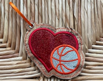 May include: A cork-backed key chain with a red heart and a silver basketball design. The heart is outlined in orange thread and the basketball is outlined in orange and white thread.