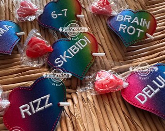 May include: Heart-shaped, rainbow-colored felt patches with embroidered text, including "RIZZ", "SKIBIDI", and "BRAIN ROT". Each patch has a white stick and is accompanied by a wrapped red candy. The patches are arranged on a woven surface.