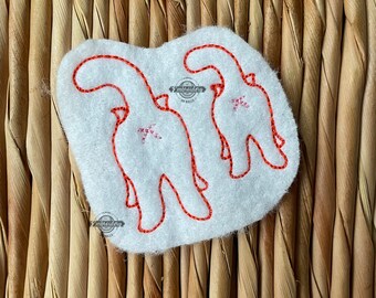 May include: White felt applique featuring two stylized dinosaur silhouettes outlined in red thread. Each dinosaur has a small pink star detail. The applique is on a woven, light brown background.