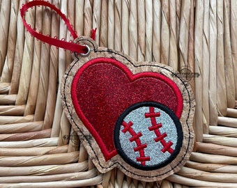 May include: A cork heart-shaped ornament with a red glitter heart and a silver baseball with red stitching. The ornament has a red ribbon loop for hanging.