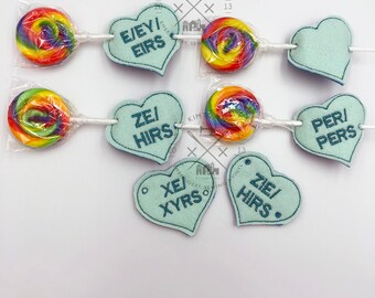 May include: A garland of rainbow lollipops and felt hearts with embroidered text. The text on the hearts reads "E/EY/EIRS", "ZEI/HIRS", "PER/PERS", "XE/XYRS", and "ZIE/HIRS".