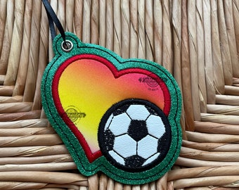 May include: A green, red, and yellow heart-shaped ornament with a black and white soccer ball inside. The ornament has a loop at the top for hanging.