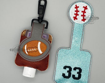 May include: Two hand sanitizer holders with a football and baseball design. The football holder is brown with a gray strap and a white football design. The baseball holder is light blue with a white baseball design and the number 33.