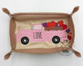 May include: A pink truck with a bed full of red, purple, and pink hearts. The truck has the word "LOVE" embroidered on the side. The truck is sitting in a gold leather tray with red stitching.