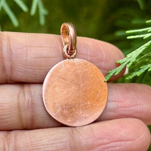 Peut inclure: Un pendentif rond de couleur cuivre avec une surface lisse légèrement texturée. Le pendentif a une boucle en haut pour l'attacher à une chaîne.