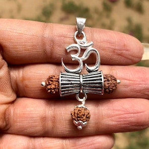 May include: Silver pendant featuring the Om symbol, a traditional Indian religious symbol, with a pair of silver damru, a small hand drum, and rudraksha beads.