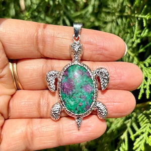 Könnte beinhalten: Silberner Schildkrötenanhänger mit einem grünen und violetten Edelstein. Der Anhänger ist mit strukturierten Silberakzenten verziert.