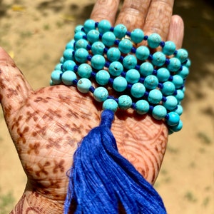 May include: A turquoise blue mala bead necklace with a blue tassel. The necklace is made of round beads and is strung on a blue cord.