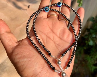 May include: Two black beaded necklaces with silver accents and blue evil eye charms. The necklaces are layered and have a silver tassel.
