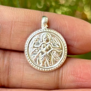 Puede incluir: Un colgante de plata con un diseño en relieve detallado. El colgante circular presenta una figura central con los brazos extendidos, rodeada por un borde de cuentas. El colgante tiene un pequeño lazo en la parte superior para una cadena.