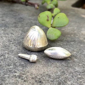 May include: Three silver, decorative, miniature,  objects. The largest object is a cone-shaped,  with a ribbed texture. The second object is a small, oval-shaped object with a ribbed texture. The smallest object is a small, cylindrical object with a rounded top.