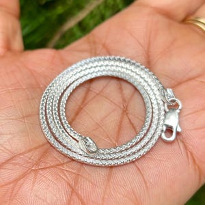 Op de afbeelding: Een opgerolde zilverkleurige slangenketting met een getextureerd, schubachtig patroon. De ketting heeft een slangenkop hanger en een sluiting. De sieraden worden op de handpalm van een persoon getoond.