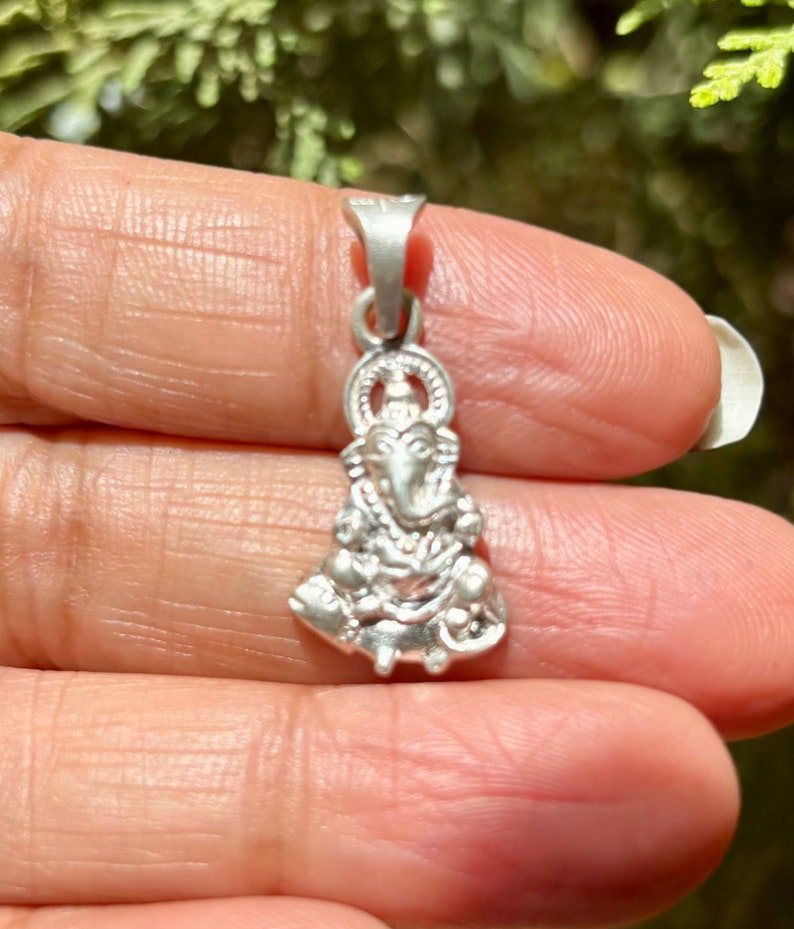 May include: Silver pendant featuring a detailed depiction of the Hindu deity Ganesha. The pendant is intricately crafted with a three-dimensional design, showcasing Ganesha's trunk, large ears, and other distinctive features.