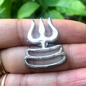 Puede incluir: Un colgante de color plateado con un diseño de tridente de tres puntas y una base circular. El tridente es un símbolo del dios hindú Shiva.