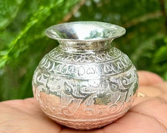 May include: A small silver decorative pot with intricate floral and heart-shaped designs. The pot has a round body and a slightly flared opening.