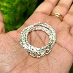 May include: A silver chain necklace with a box chain design. The chain is coiled in a circle and held in a hand.