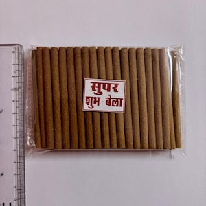 May include: A packet of brown incense sticks, neatly arranged and sealed in clear plastic. A small, rectangular label with text in a foreign language is visible. A ruler is placed on the left side of the image.