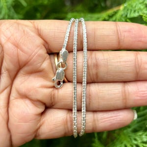 May include: A silver chain necklace with a box chain design. The chain is shown in a hand, with the clasp visible.