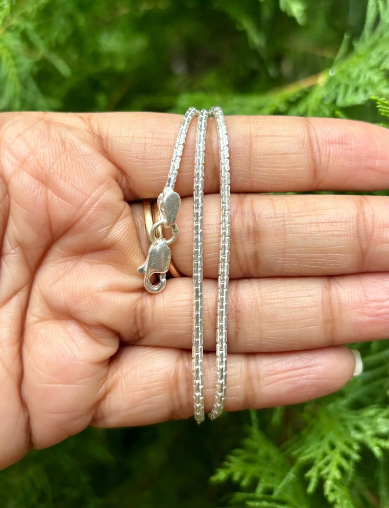 May include: A silver chain necklace with a box chain design. The chain is shown in a hand, with the clasp visible.