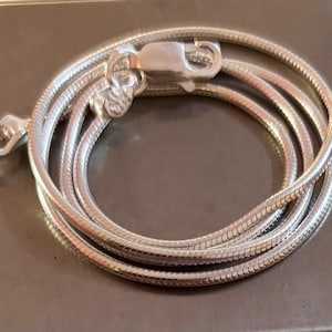 Puede incluir: Un collar de cadena de serpiente plateada enrollada con un cierre de mosquetón. La cadena está hecha de pequeños eslabones interconectados, creando una textura suave y flexible. El cierre es un diseño simple y seguro.