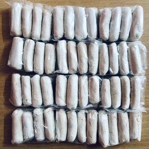 May include: A close-up of a large quantity of white, cylindrical clay bars, individually wrapped in clear plastic, arranged in rows on a wooden surface.