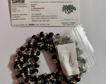 May include: A black beaded necklace with a silver chain. The necklace is 109 beads and approximately 6 mm in diameter. The necklace is accompanied by a certificate of authenticity from VEGGA GEMS TESTINGS LABS. The certificate states that the necklace is made of natural Karungali wood, also known as Ebony Wood.
