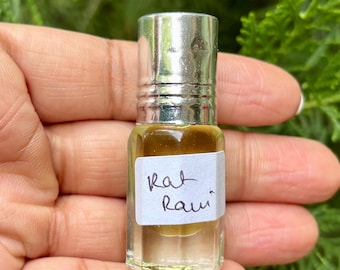 May include: A small, clear glass bottle with a silver cap, filled with a dark amber liquid. A white label with the handwritten text "Rat Rami" is affixed to the bottle. The bottle is held in a hand against a backdrop of green foliage.