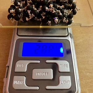 May include: A digital scale with a blue display showing 28.8 grams. The scale has buttons labeled "PCS", "UNITS", "ON/OFF", "TARE", and "LIGHT". A pile of black beads with silver accents is on the scale.