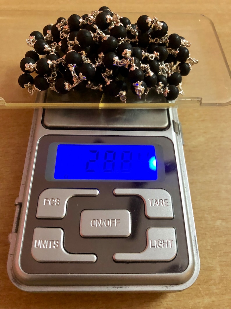 May include: A digital scale with a blue display showing 28.8 grams. The scale has buttons labeled "PCS", "UNITS", "ON/OFF", "TARE", and "LIGHT". A pile of black beads with silver accents is on the scale.